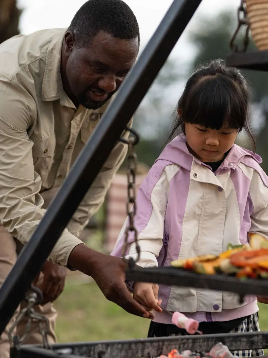 A camp cook lifting a grill of roasted vegetables while a small girl in a pink jacket watches from beside him