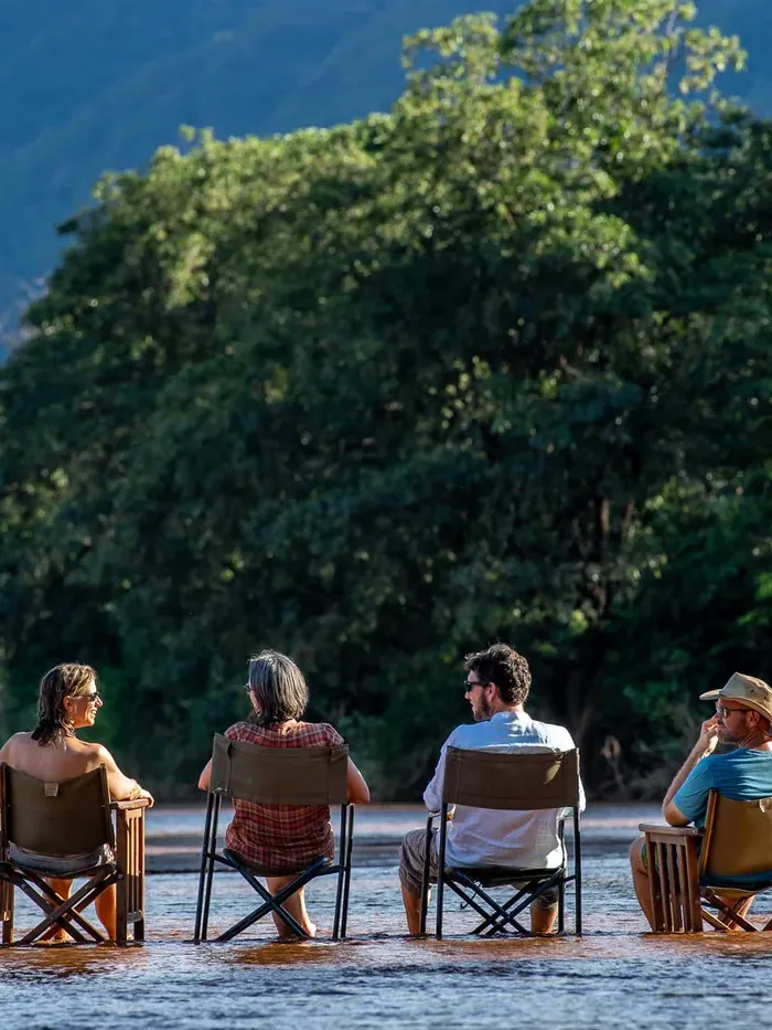 Four friends seen from behind, sitting in camp chairs set in the shallows of a river below a wooded hillside