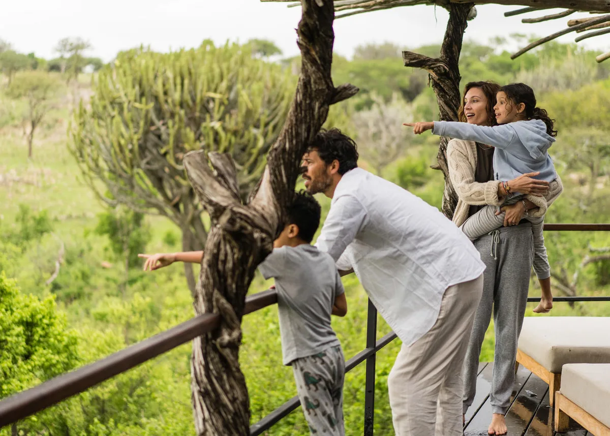 A family of four leaning on a lodge railing, the parents and children pointing out something in the bush below