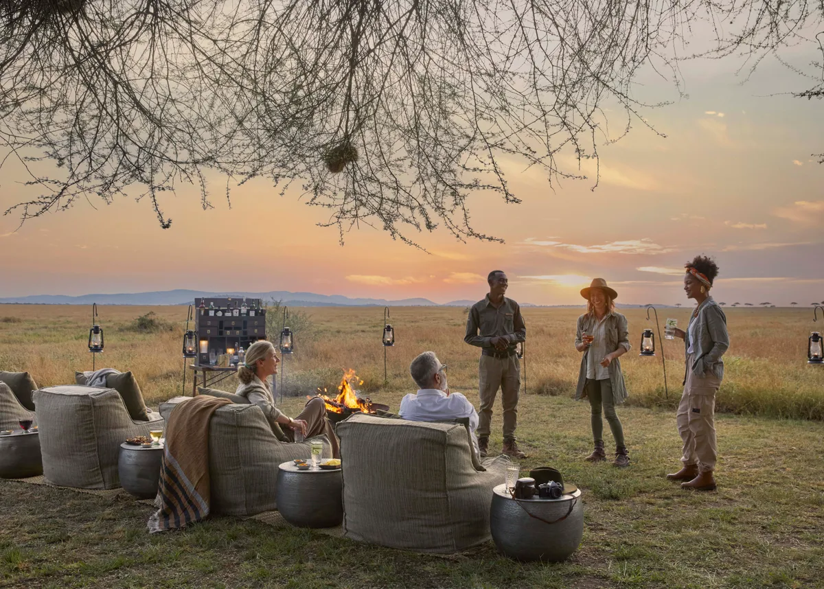 Guests and their guides around a fire pit on the open plains, drinks in hand as the sun sets