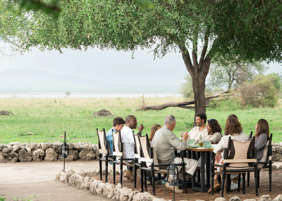 A family at a long lunch table set in the shade of an acacia, with the lake and hills beyond