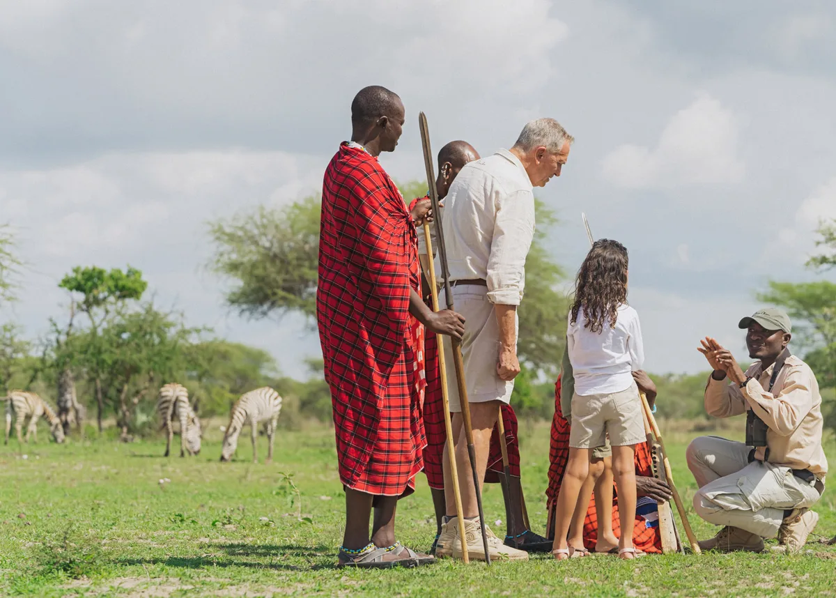 A Maasai guide leading a family on a walking safari, a herd of zebra grazing in the grass behind them