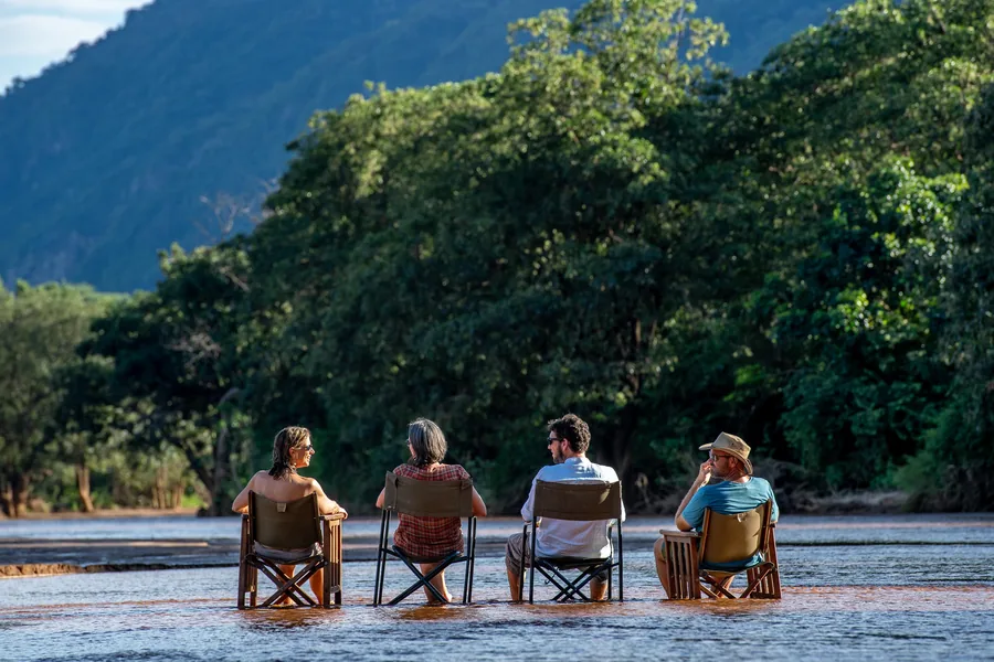 Four guests sitting in camp chairs set out in the shallows of a river running high and silty, dense green forest and a hillside rising behind them