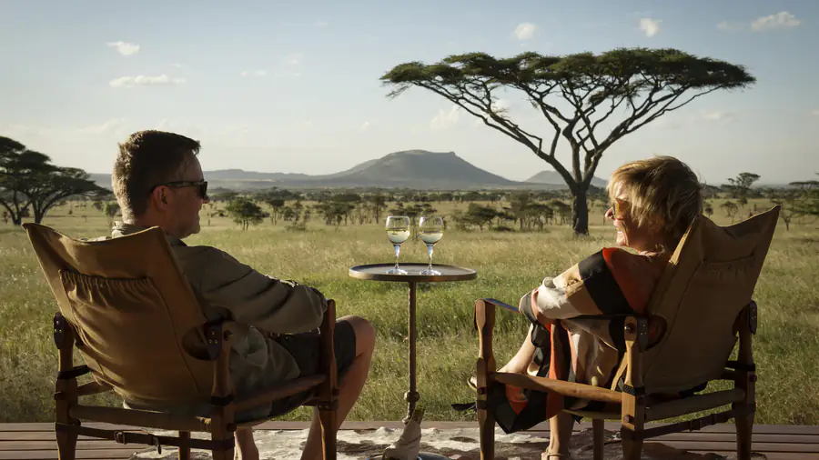 Two guests in safari chairs on a camp deck, glasses of wine on a table between them, looking out at an acacia and a flat-topped hill across the grass