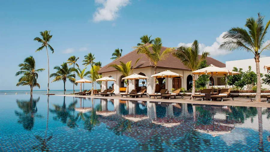 An infinity pool lined with palms and white parasols in front of a thatched beach lodge, the sea beyond it and the palms reflected in the water