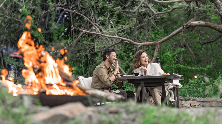 A couple laughing over dinner at a table laid out in the bush, a fire burning in the foreground and lanterns hung in the trees behind them