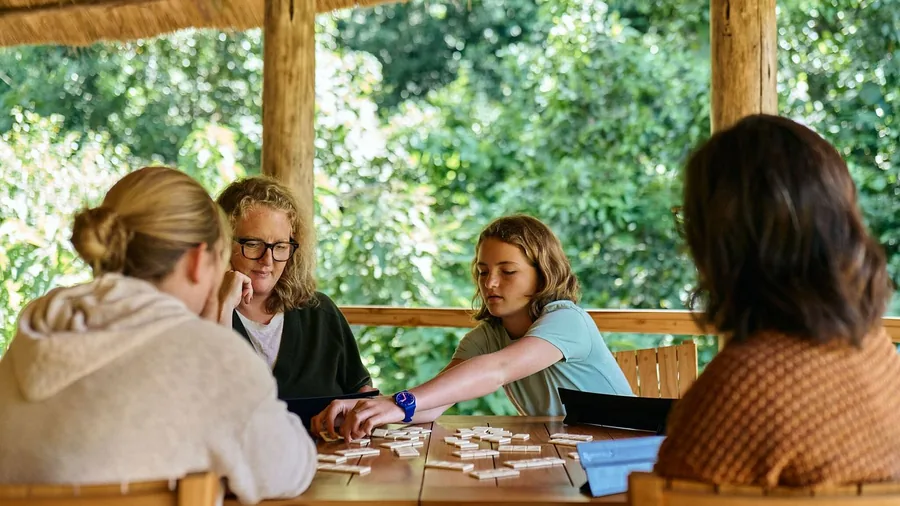 Four people around a wooden table under a thatched roof, mid-game with tiles laid out between them and forest behind
