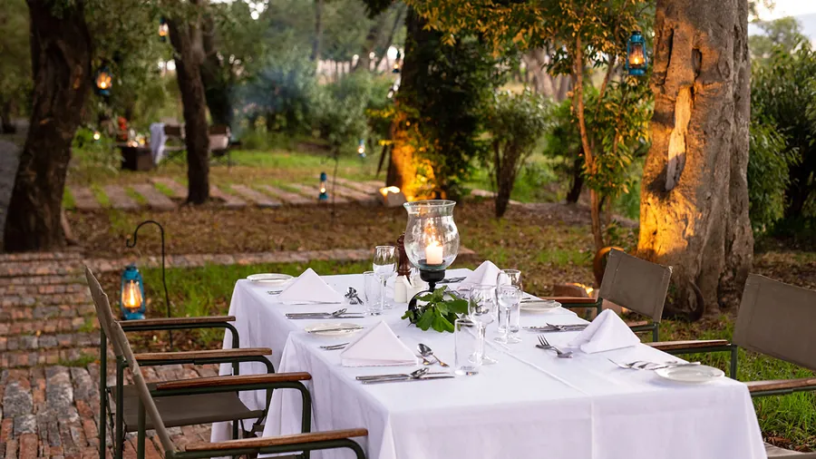A table laid with white linen and a candle in a hurricane glass on a brick terrace, uplit trees and lanterns in the garden behind