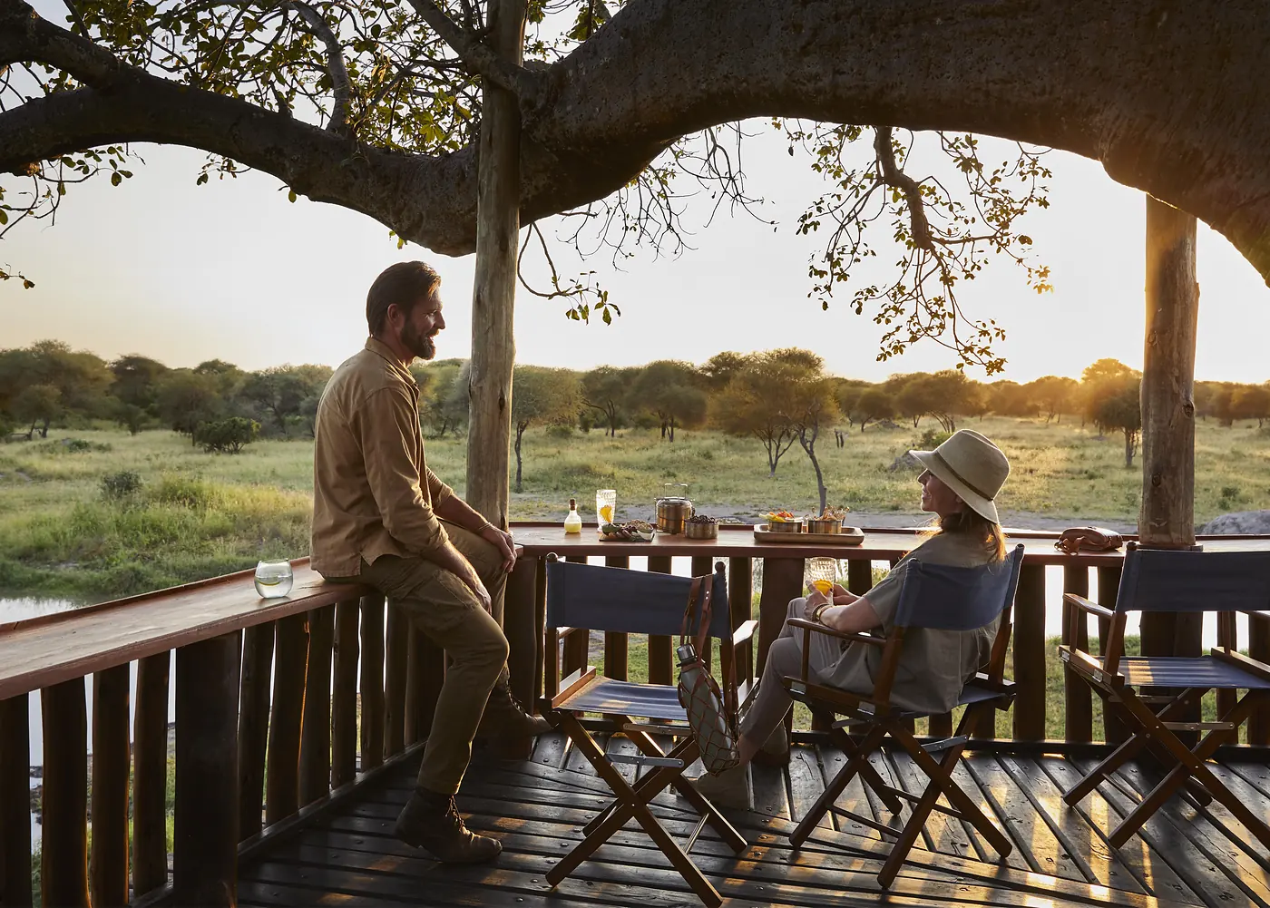 A couple taking breakfast on a lodge deck at sunrise, the plains opening out below them