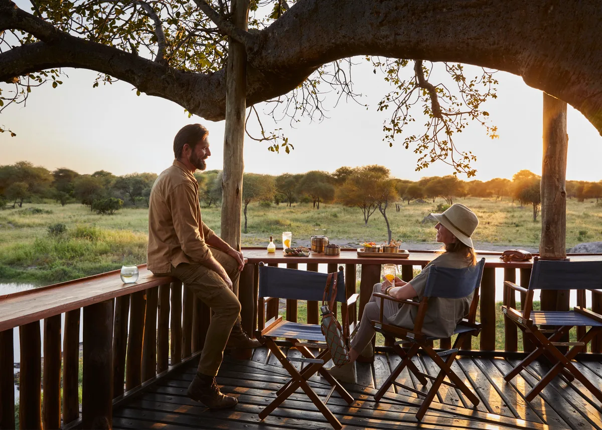 A couple taking breakfast on a lodge deck at sunrise, the plains opening out below them