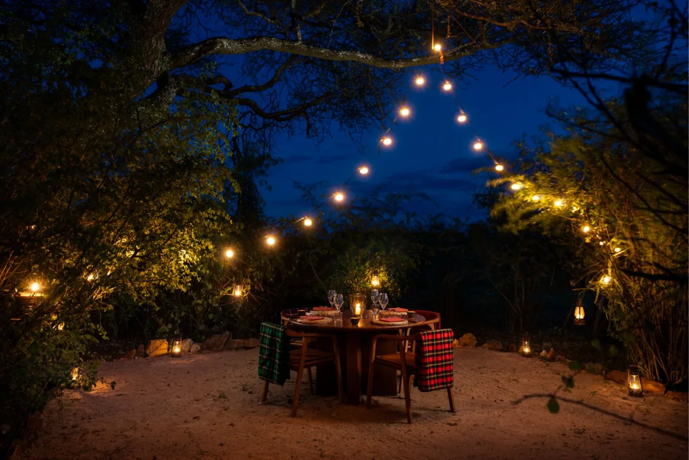 A table laid for two in a clearing after dark, festoon lights strung through the trees above it and lanterns set out around the sand