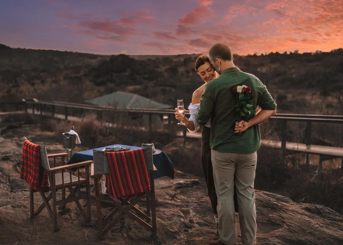 A couple standing arm in arm with champagne at a private sundowner table on a rocky outcrop, the sky turning red behind them