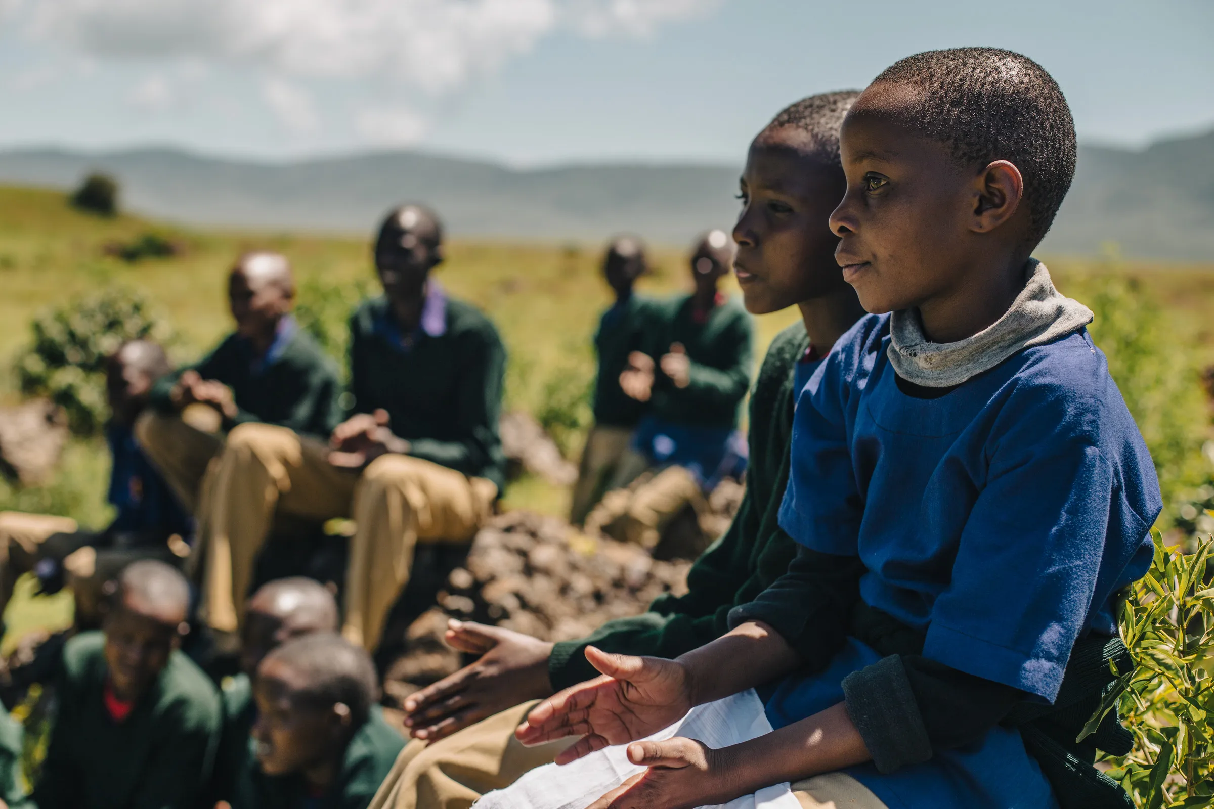 Schoolchildren in uniform sitting together on a grassy hillside, listening, with the highlands behind them