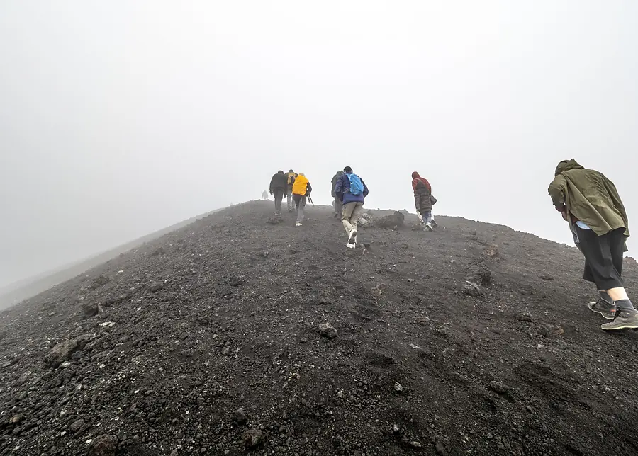 A file of trekkers working up a ridge of black volcanic ash in thick cloud, high on the mountain