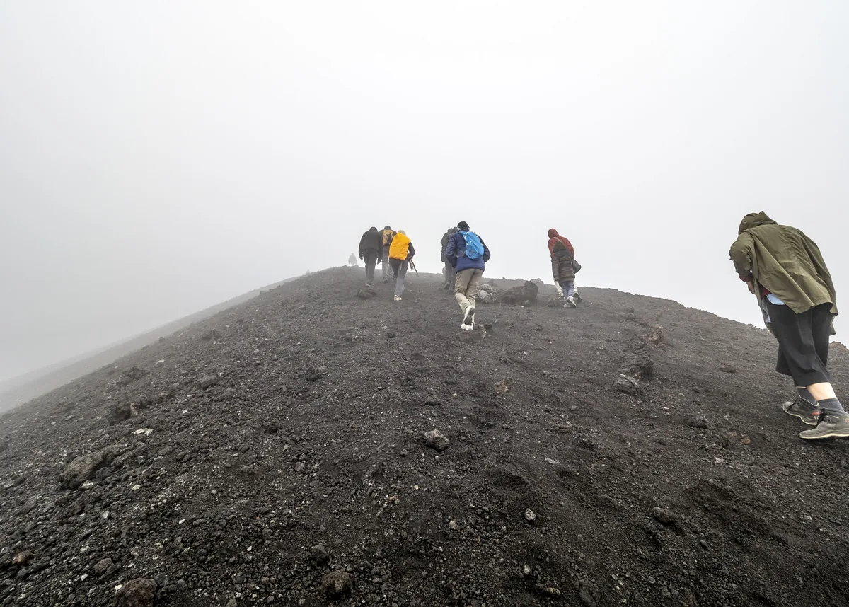 A file of trekkers working up a ridge of black volcanic ash in thick cloud, high on the mountain