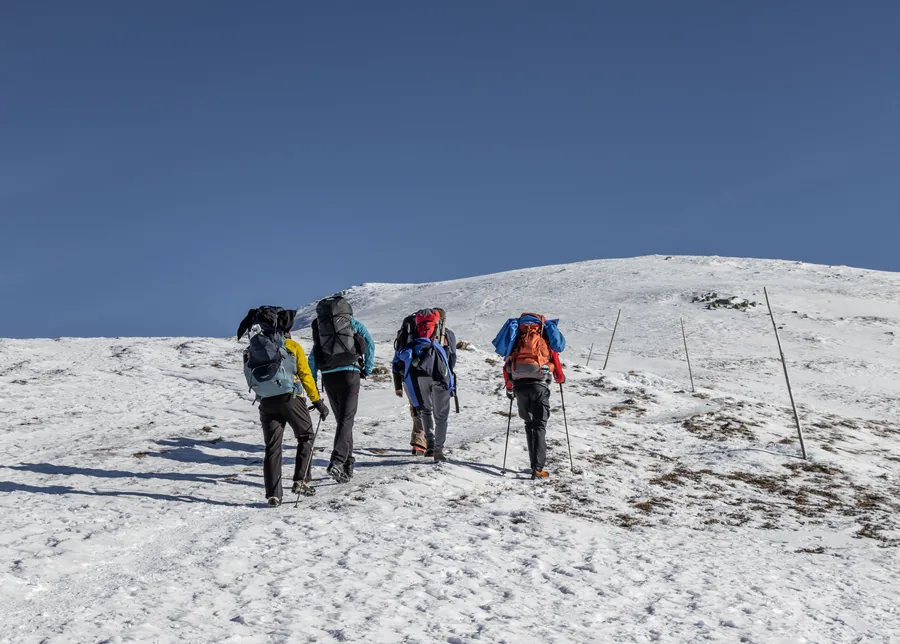 Four trekkers in down jackets walking up a snowfield in single file with poles and full packs, the rounded summit ahead of them under a clear sky