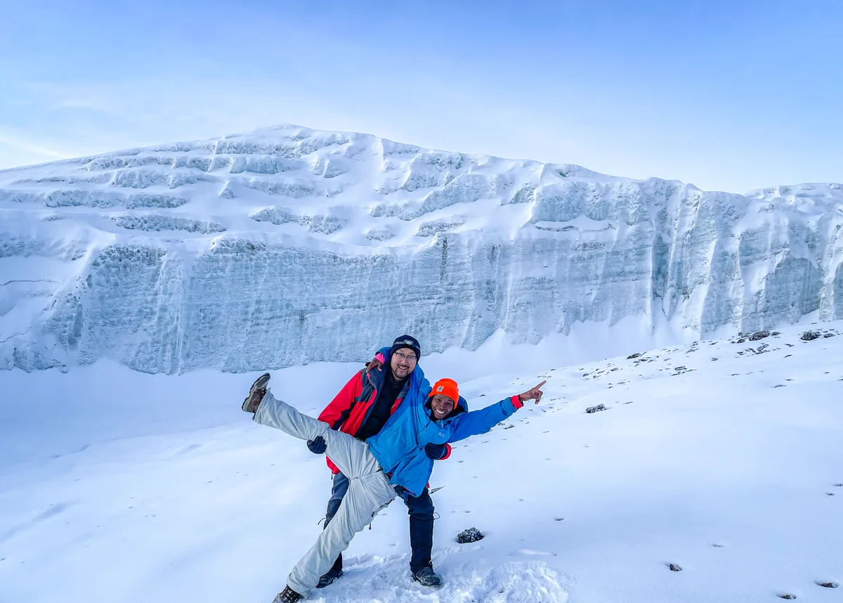 Two climbers celebrating in the snow at the top of Kilimanjaro, the summit glacier wall rising behind them