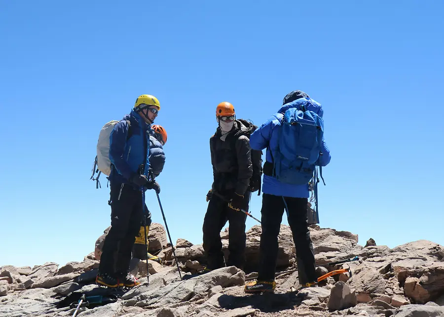 Three climbers in cold-weather kit stopped on bare rock high on the mountain under a deep blue sky