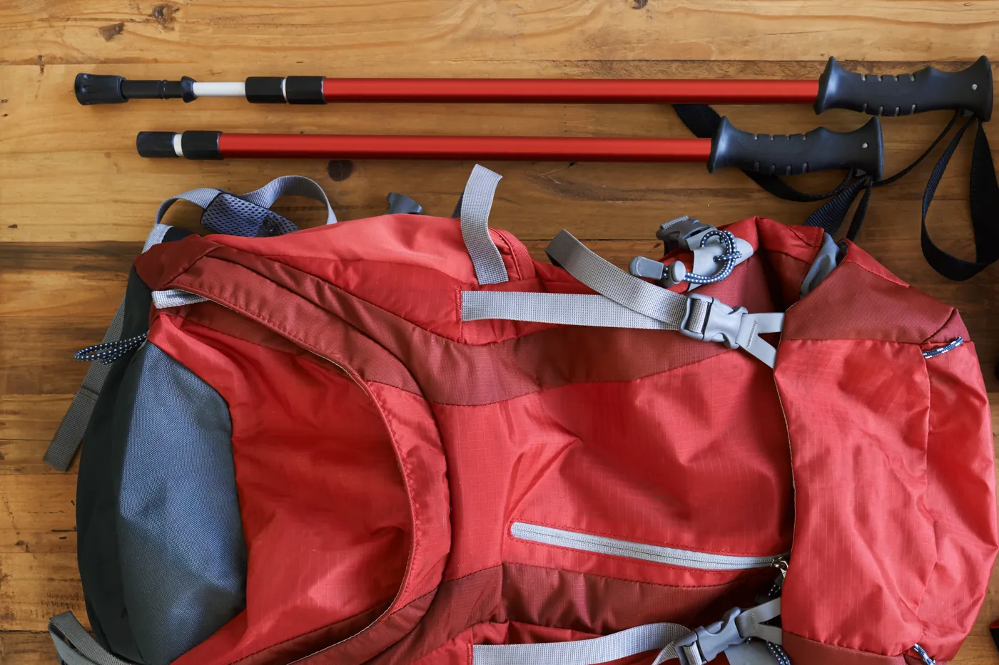 A red framed rucksack lying on floorboards with a pair of telescopic walking poles above it