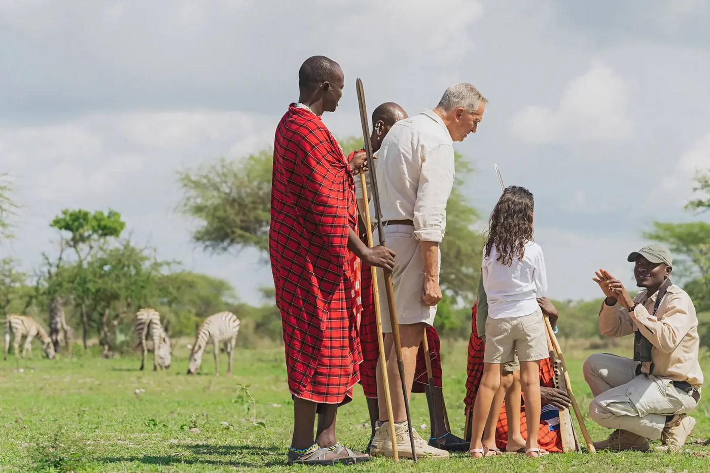 A Maasai guide in a red shuka leaning on his staff beside a family on a bush walk, zebra grazing on the plain behind them