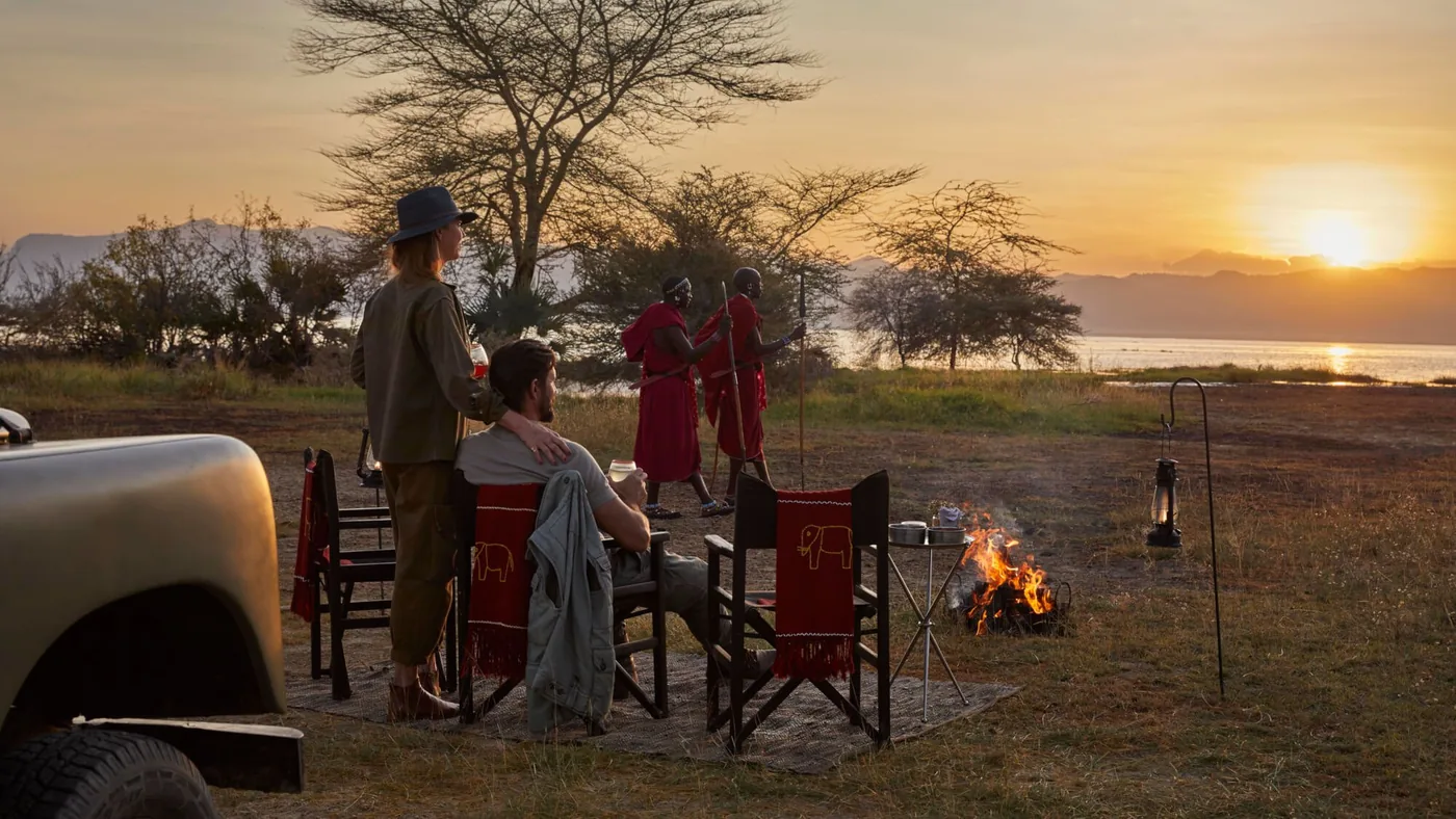 Guests in camp chairs around a fire at a sundowner on the shore of Lake Manyara, two Maasai hosts standing with them and the sun going down over the water