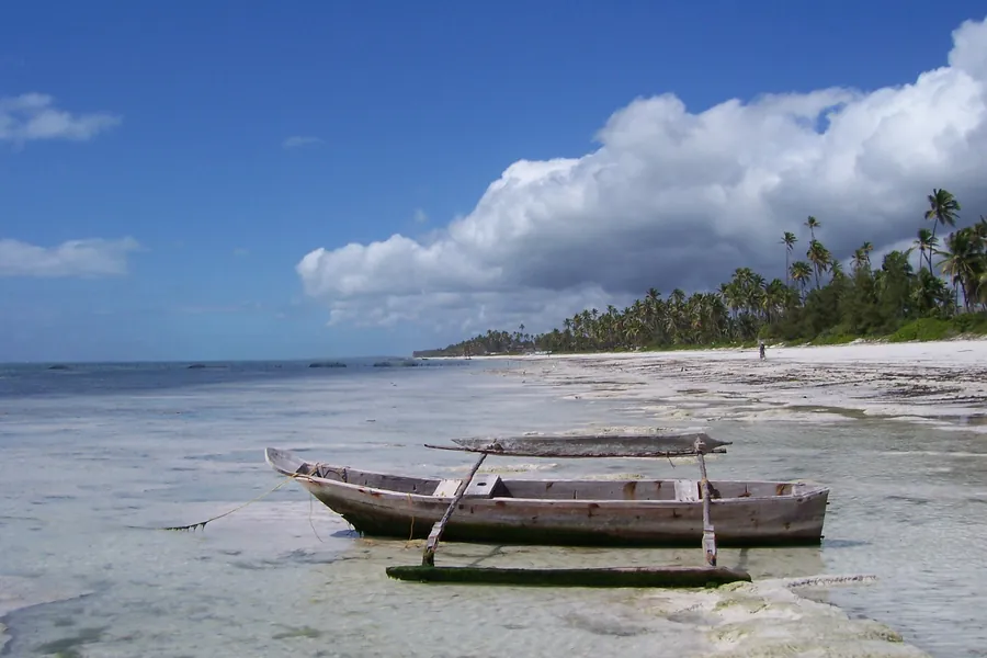 A wooden outrigger canoe resting in the low-tide shallows on the east coast near Bwejuu, with a palm-lined beach behind