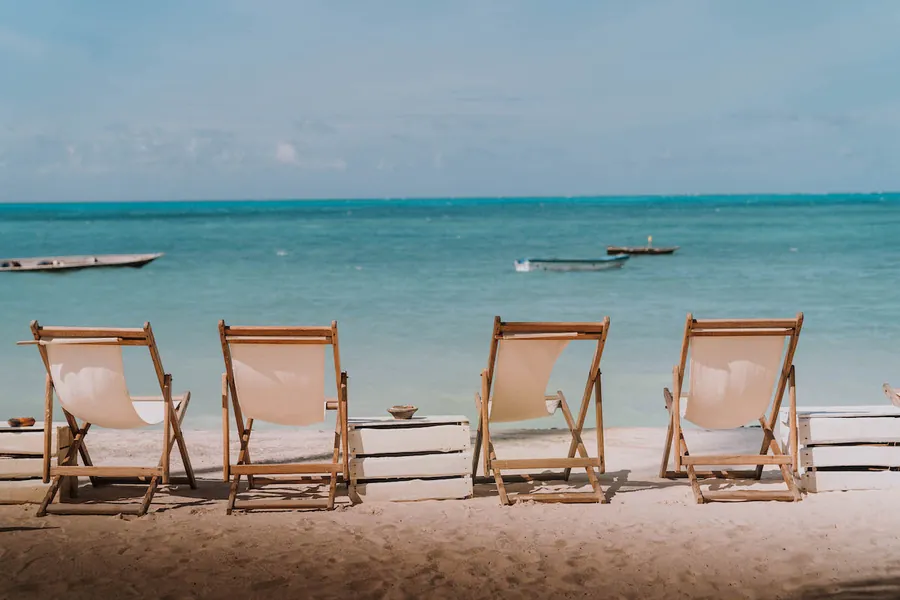 Four canvas deck chairs on the sand at Jambiani facing turquoise water, wooden fishing boats moored offshore